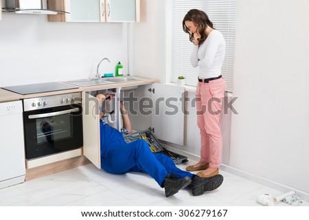 Young Repairman Repairing Sink Pipe While Woman Standing In The Kitchen