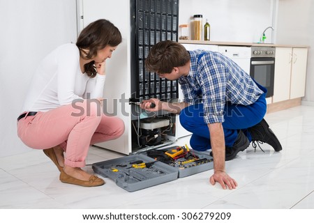 Young Repairman Repairing Refrigerator In Front Of Beautiful Woman