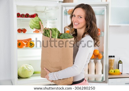 Happy Woman Holding Grocery Shopping Bag With Vegetables In Front Of Open Refrigerator