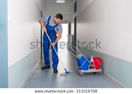Full length of young male worker with broom cleaning office corridor