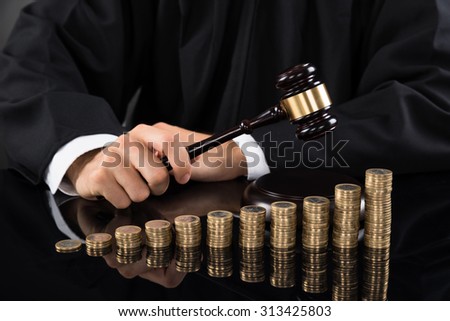 Close-up Of Judge Hands Hitting Gavel In Front Of Stacked Coins At Desk