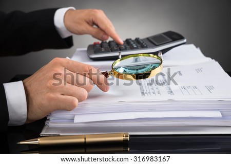Close-up Of Businessperson Hands Checking Invoice With Magnifying Glass At Desk