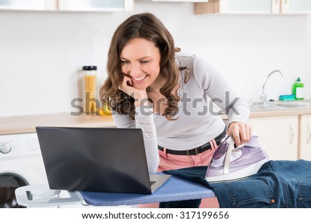 Happy Woman Looking At Laptop While Ironing Cloth On Ironing Board