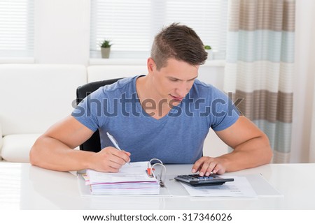 Young Man Calculating Invoice With Calculator At Desk