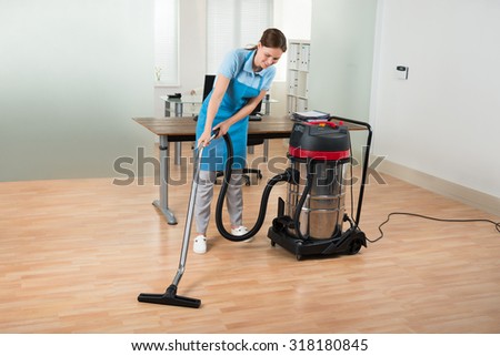 Happy Female Worker Cleaning Floor With Vacuum Cleaner In Office