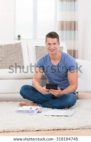 Young Man Sitting On Carpet Calculating Budget With Calculator