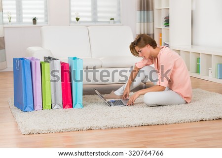 Young Woman Sitting On Carpet With Laptop And Shopping Bags In Living Room