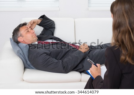 Patient Relaxing On Couch In Front Of A Female Psychiatrist With Clipboard