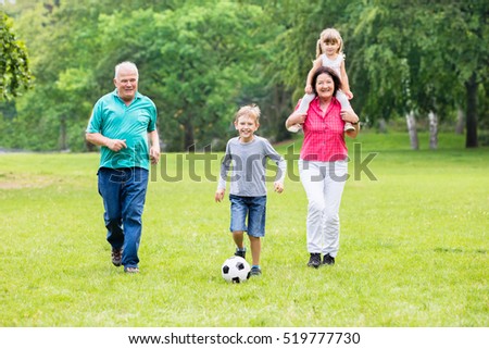Happy Family Playing Soccer Game With Grandchildren Together In Park. Running For Ball