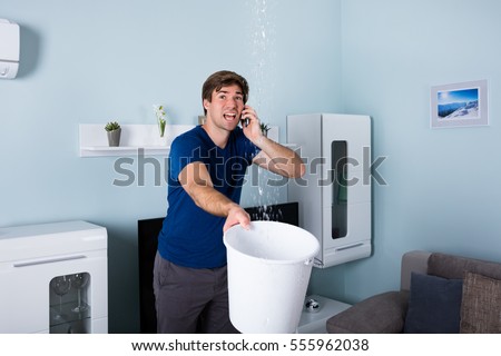 Worried Man Calling Plumber While Leakage Water Falling Into Bucket At Home