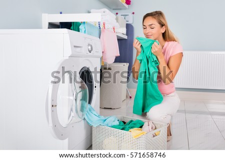 Young Smiling Woman Smelling Clothes After Washing In Washing Machine At Utility Room