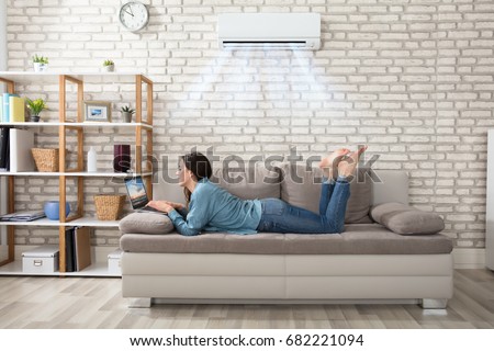 Woman Lying On Sofa Using Laptop Enjoying The Cooling Of Air Conditioner At Home