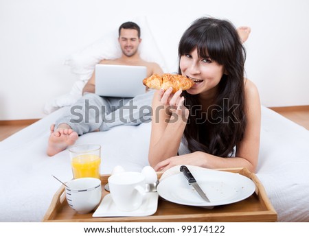 Young woman eating breakfast in bed while man using laptop