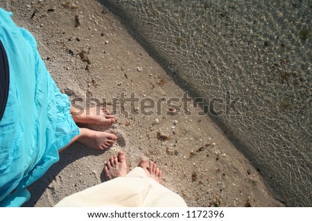 Lovers feet together on a deserted beach with the ocean lapping at their toes.