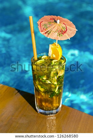 Glass of ice cold cocktail mojito with umbrella near swimming pool