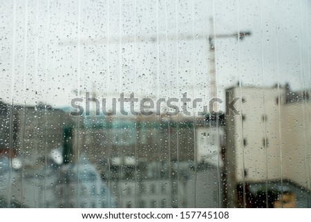 Silhouettes of the building cranes in city behind the wet glass, where on the street cloudy and rain