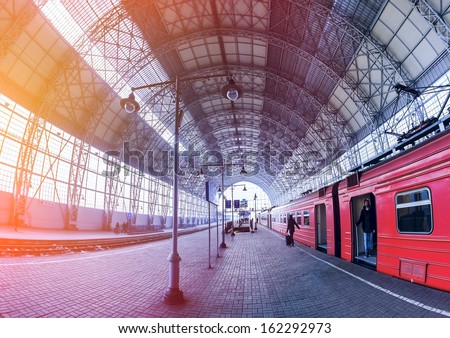 Covered railway station with train and silhouettes of hurrying people. Young man in the tambour train