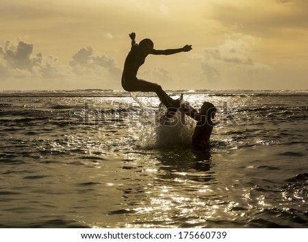 Silhouette of young girl jumping out of the ocean, which throws strong two man on the background of the expiring sunset