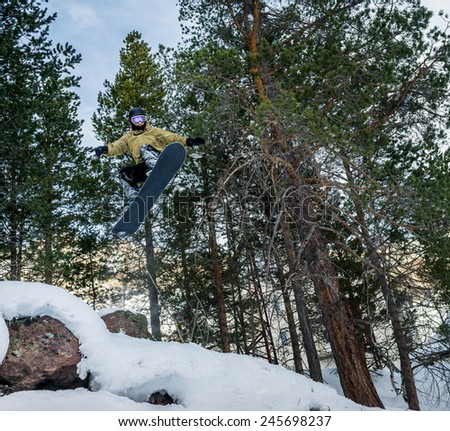 Snowboarder jumping over the snow-covered stone on the background forest. Low angle view.