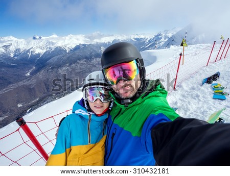 Happy couple snowboarders standing on edge of mountain peaks and taking selfie portrait with camera or smartphone on  background of snowy mountains in ski resort
