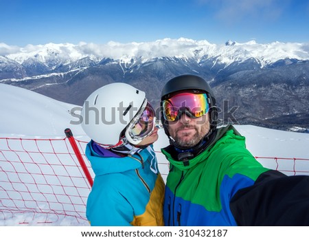 Happy couple snowboarders standing on edge of mountain peaks and taking selfie portrait with camera or smartphone on  background of snowy mountains in ski resort