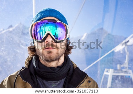 Portrait of bearded young man in sunglass mask, rises to the top in the cabin cable car at the ski resort on the background of mountains and blue sky