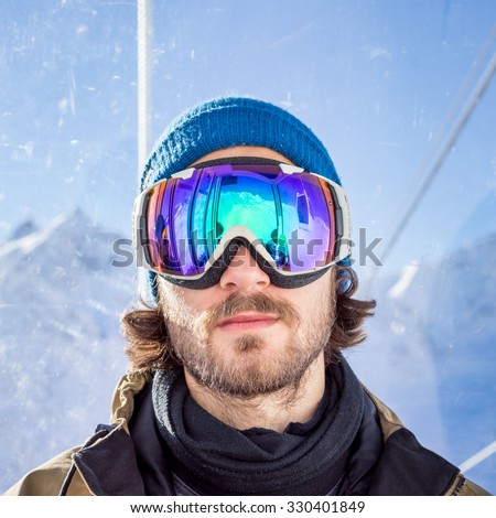 Portrait of bearded young man in sunglass mask, rises to the top in the cabin cable car at the ski resort on the background of mountains and blue sky
