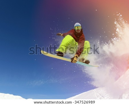 Jumping snowboarder keeps one hand on snowboard in mountains in ski resort on blue sky background