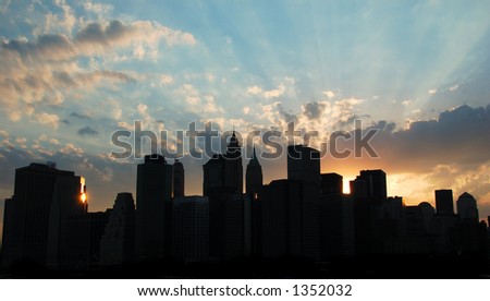 Downtown Manhattan Skyline with sunset.  There are rays of light emerging from behind the buildings.  The buildings are silhouetted with a small amount of shadow detail.  There are clouds  & blue sky.