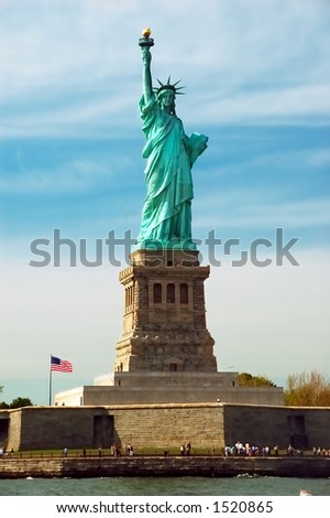 The Statue of Liberty on a sunny day with the American flag waving.  There is a blue sky with wispy clouds.  The torch is nicely lit in the sunlight.  There are visitors visible at the base.