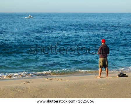 A man is surf fishing from a beach.  The ocean is a brilliant blue and choppy.  There is a small amount of surf.  He is wearing a red cap and is barefoot.