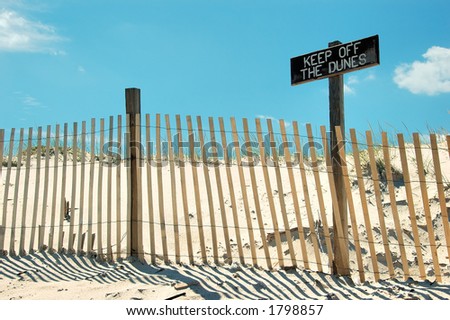 Beach scene with fence and sign that says keep of the dunes.  The sign is weathered.