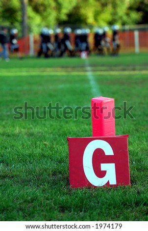 Goal Line sign for American Football.  A team of players is visible in the blurred background.  There is green grass and dappled sunshine.