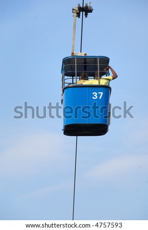 Blue Cable Car from a sky ride high above a theme park.  Two people, a man and a woman are visible.  There is a blue sky with thin clouds.