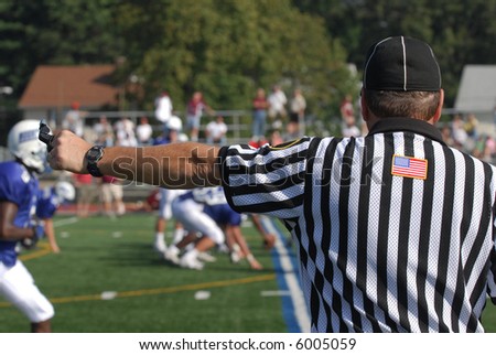 A referee at an North American Football game.  He has his arm extended his whistle and stop watch are visible.  There is an active game with players and spectators in the blurred background