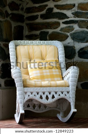 Old style wicker rocking chair with yellow cushions on porch with stone wall in background