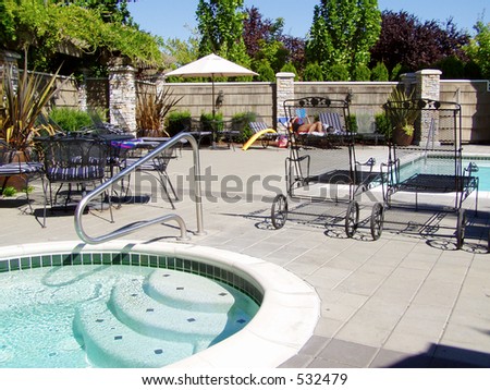 Hot tub pool in left foreground on sunny day with black iron lounge chairs, tables, and larger pool at back.