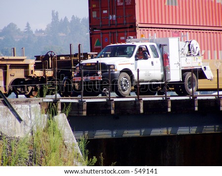 Train track worker drives special tool truck on tracks over tunnel with freight train in background.