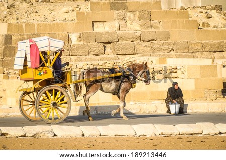 GIZA, EGYPT - NOVEMBER 19, 2011: People in a horse cart ride past a woman selling water at the Great Pyramids Site in Giza, Egypt. Giza is the third largest city in Egypt, neighboring with Cairo.