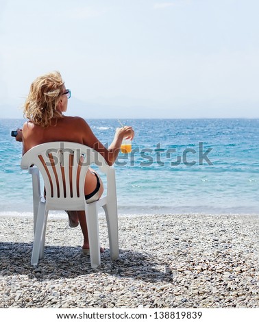 girl, blonde sits in a chaise lounge drinks cold orange juice and looks at the turquoise sea. Clear sunny summer day, mountains on a background. Balkans, Greece, Europe