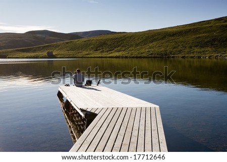 man with notebook sitting on pier above summer lake