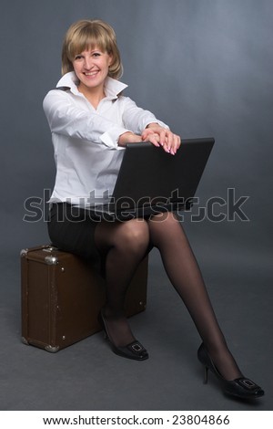 young business woman in white shirt and black skirt sitting on suitcase with notebook on her knees on grey background