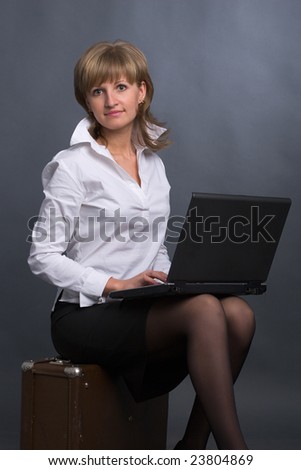 beautiful girl in white blouse and  black skirt sits on suitcase with laptop on her knees on grey background