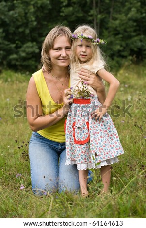 young mother and little daughter in wild flowers wreath on green meadow