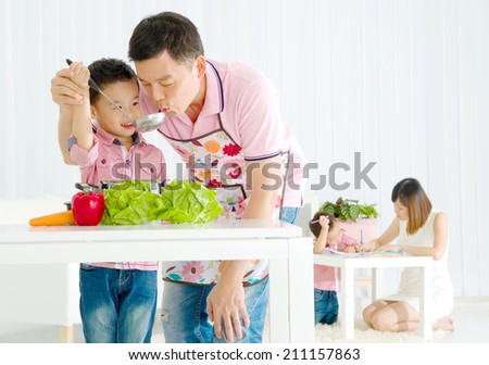 Asian father and elder son are cooking in the kitchen while mother is teaching younger son doing schoolwork.