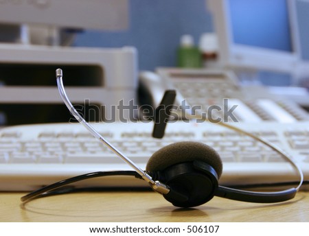 A telephone headset with help-desk equipment in the background.