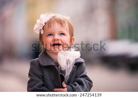 portrait of little baby girl in gray coat on the street in old city with funny smile. it is a one year old girl dressed on grey coat with big white flower in her hear and big white bow around her neck