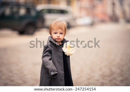 A little baby girl in gray coat stands on the street in old city. She has a big white flower brooch on her coat. Her face looks very funny with big lips and inflated cheeks