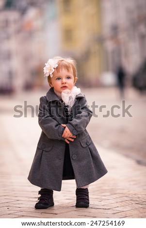 little baby stand straight on the street. one year old girl dressed on grey coat with big white flower in hear and big white bow around neck. She put hands together and smiles. background diffused