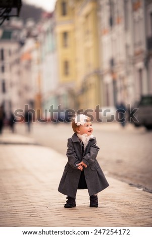 A little lady stand on the street. This is a one year old girl dressed on grey coat and white tights with big white flower in her hear and big white bow around her neck. The background is diffused